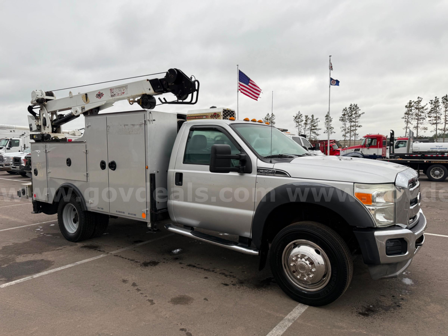 Ford F-550 service utility truck with crane/boom on a flatbed cabinet body, side compartments, white cab, silver body, American flag and equipment yard in background.