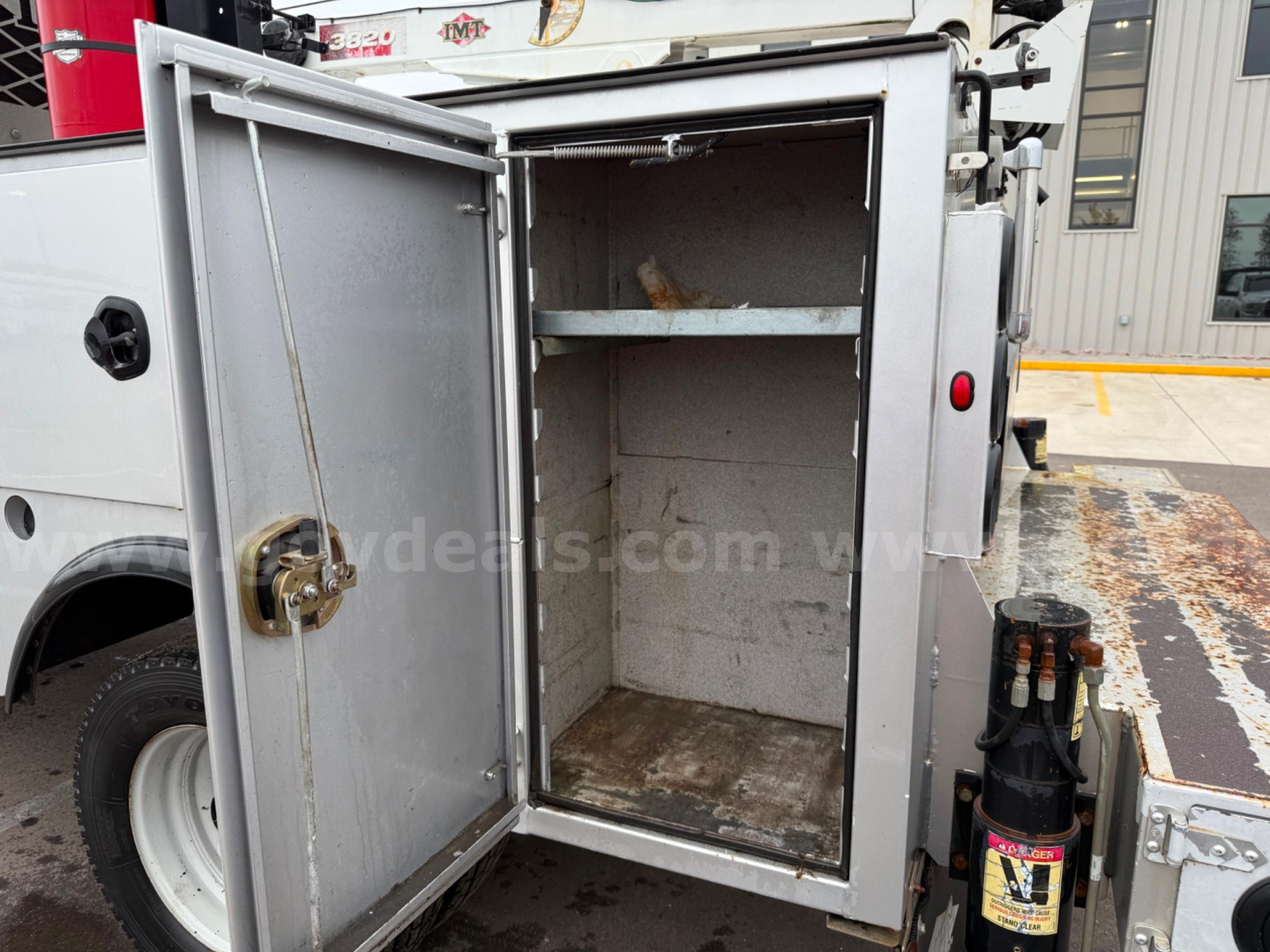 Service/utility truck rear cargo area with empty metal storage compartment and open door, showing interior shelves and weathered floor.