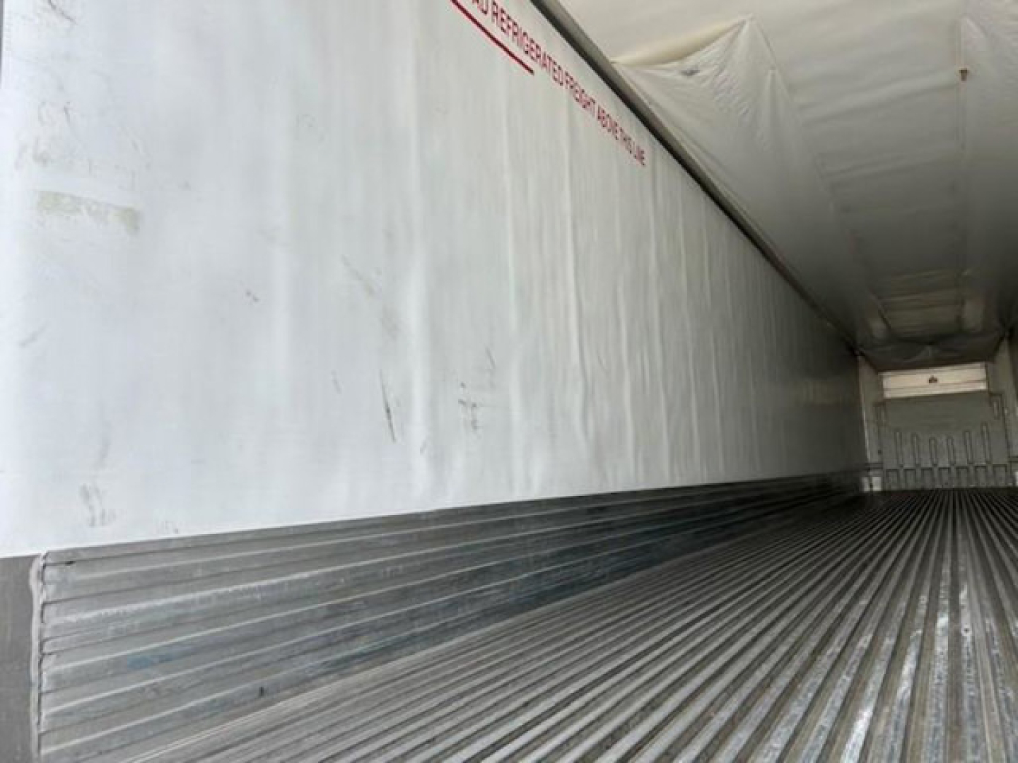 Trailer interior with metal ribbed floor and insulated walls; faint red text along top edge reading warning or instruction.