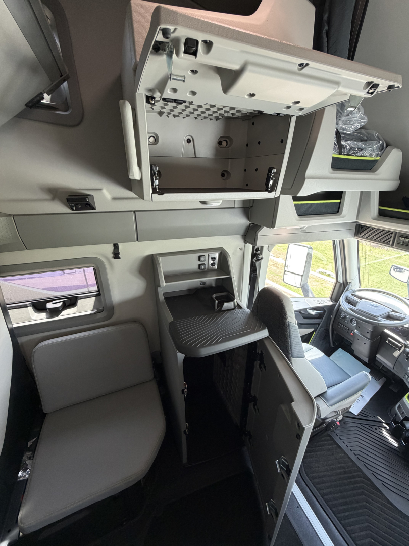Truck cab interior showing overhead bunk area with open storage cabinet and control panel in Volvo VNL cabin. Includes driver seat, dashboard, and entry area.