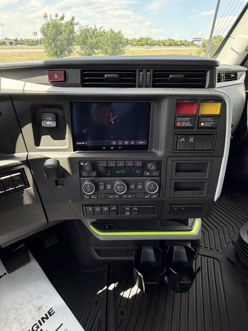 Dashboard of a Volvo VNL truck cab showing center console with touchscreen display and climate controls, hazard button, two vertical storage bays, cup holders, and yellow-green accents.