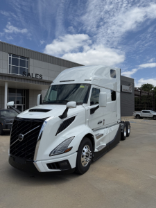 Volvo VNL64T860 truck in white, front-left view, with sleeper cab and extended mirrors, parked outside dealership. Visible text on truck panels but no legible serials.