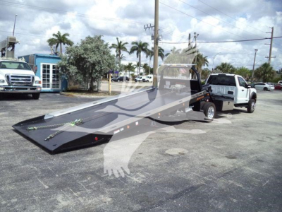 Ford F-550 rollback tow truck with a long hydraulic flatbed in a parking lot, rear view showing loading ramp and wheel-lift underframe, front cab visible in background.