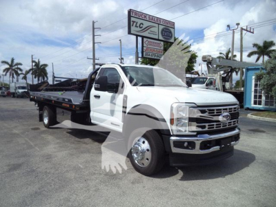 Ford F-550 rollback truck with white cab and flatbed, visible signage in background, roadside dealership lot.