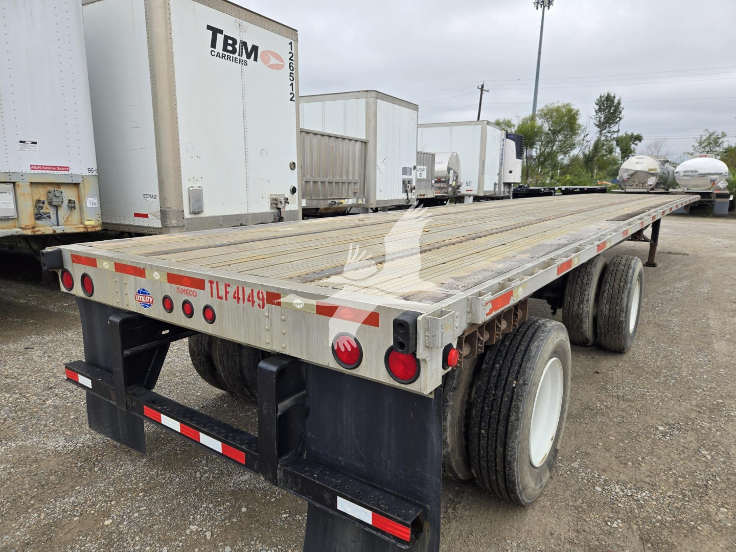 Flatbed trailer rear view showing dual axles, wooden deck, and taillights. Visible: TBMC trailer stickers, red/white reflective tape, and license plate area with lighting.