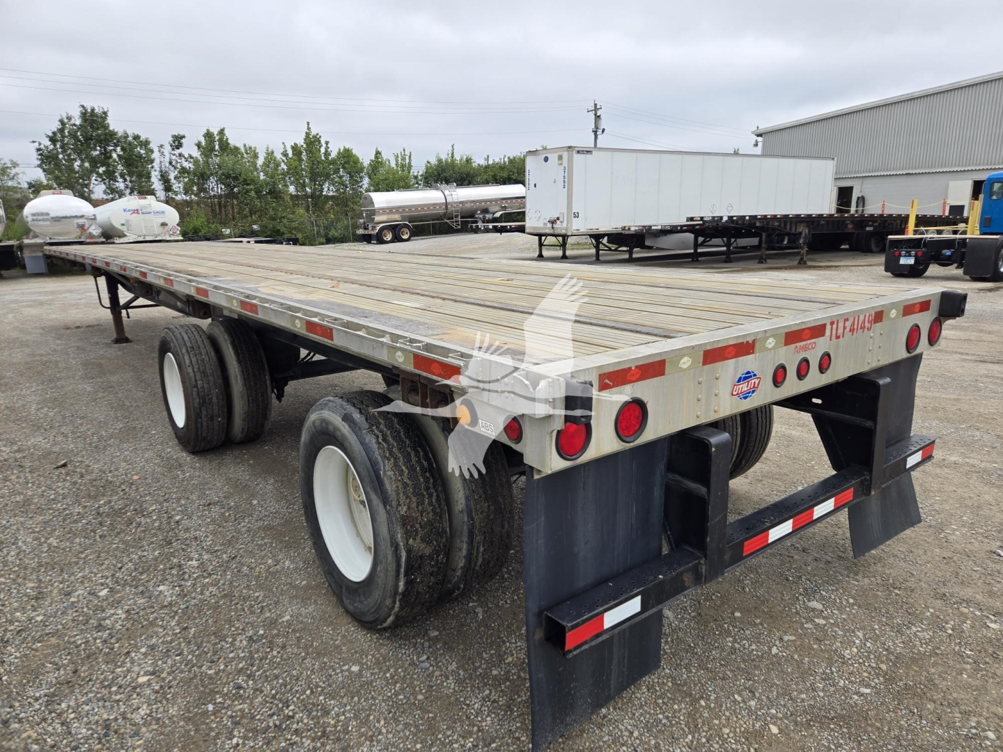 Flatbed trailer showing dual rear axles with wooden deck and rear mud flaps; reflective tape and taillights along side; stock trailer accents visible.