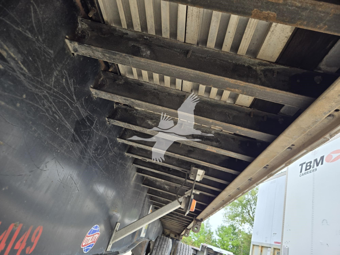 Trailer undercarriage showing flatbed support beams and underside with framing and a hanging hook.