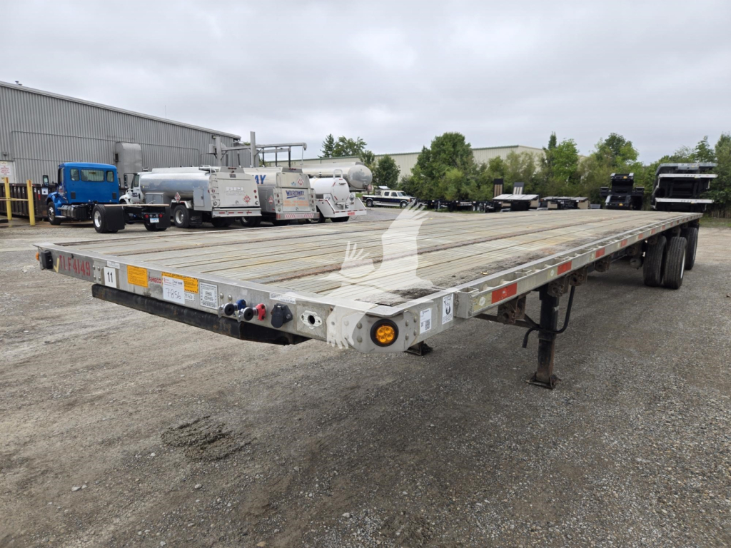 Flatbed trailer with long wooden deck showing rear lights and support legs; visible text includes stock decals and warning labels along the edge.