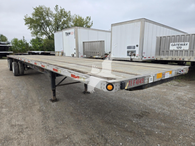 Flatbed trailer with dual rear axles and long metal deck, parking lot background with other trailers.