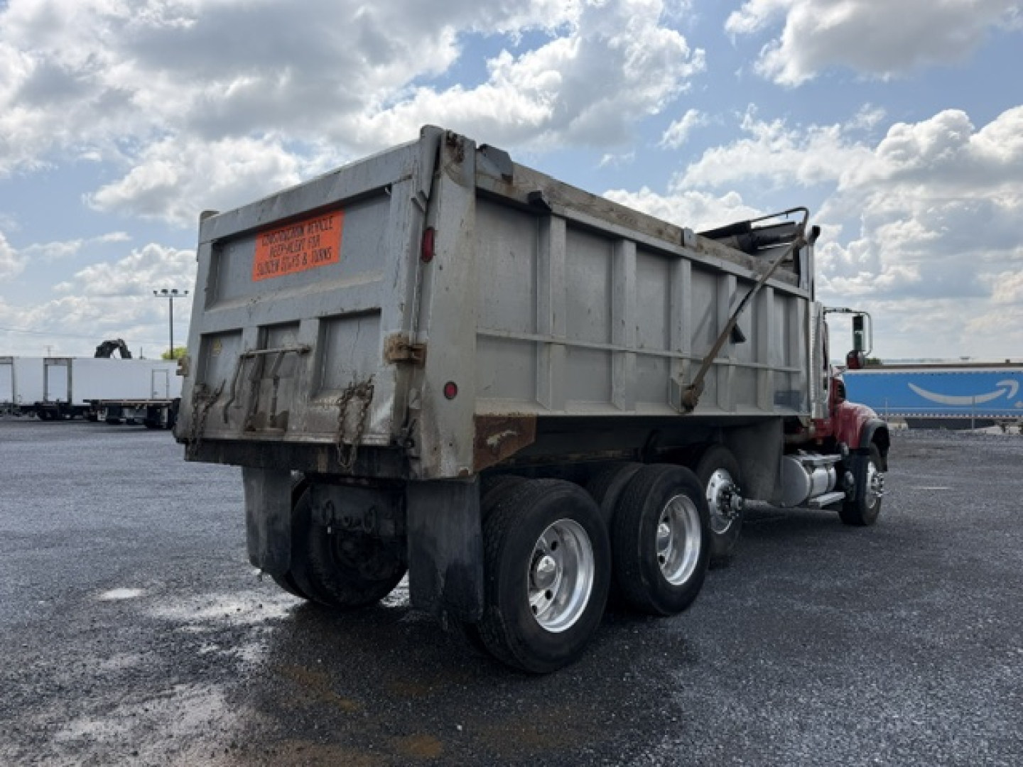 Dump truck rear view showing heavy-duty Mack CV713 dump bed and triple rear axle with undercarriage and taillight, orange warning label on tailgate.