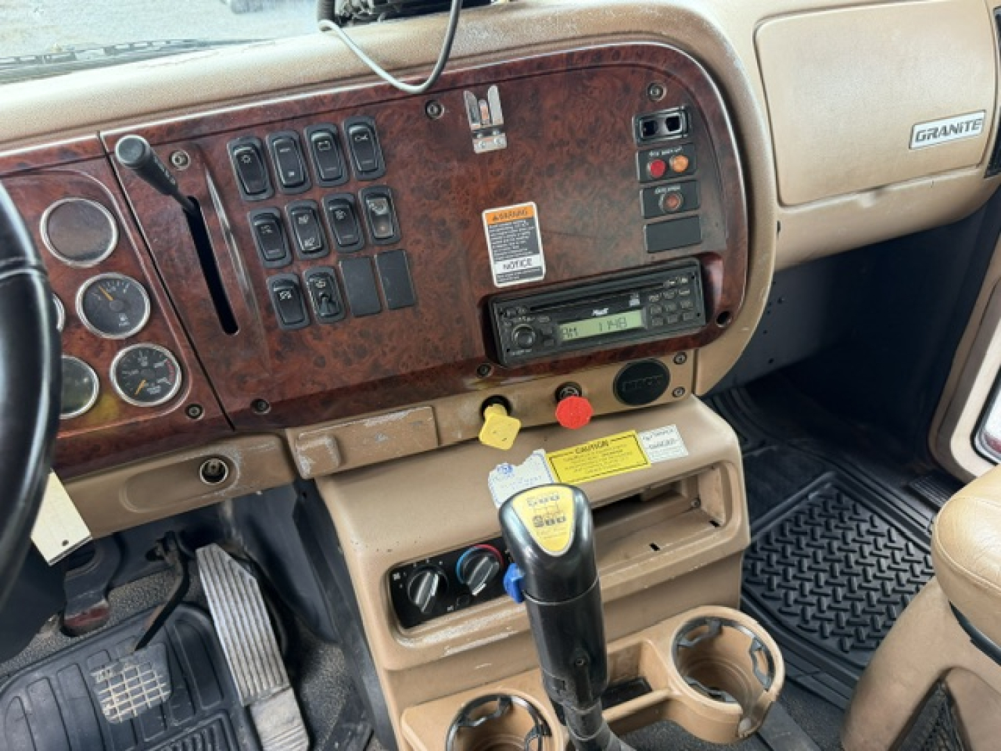 Mack CV713 dash and center console showing cluster of switches, tan interior, gear shifter, and dashboard with wood grain panel.