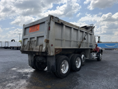 Dump truck rear view showing heavy-duty Mack CV713 dump bed and triple rear axle with undercarriage and taillight, orange warning label on tailgate.