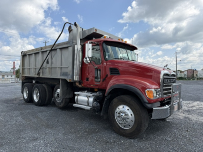 Dump truck: red Mack CV713 cab with silver dump bed, three-axle setup, front fender chrome bumper, side mirrors. Visible text on truck cab badges and mirrors.