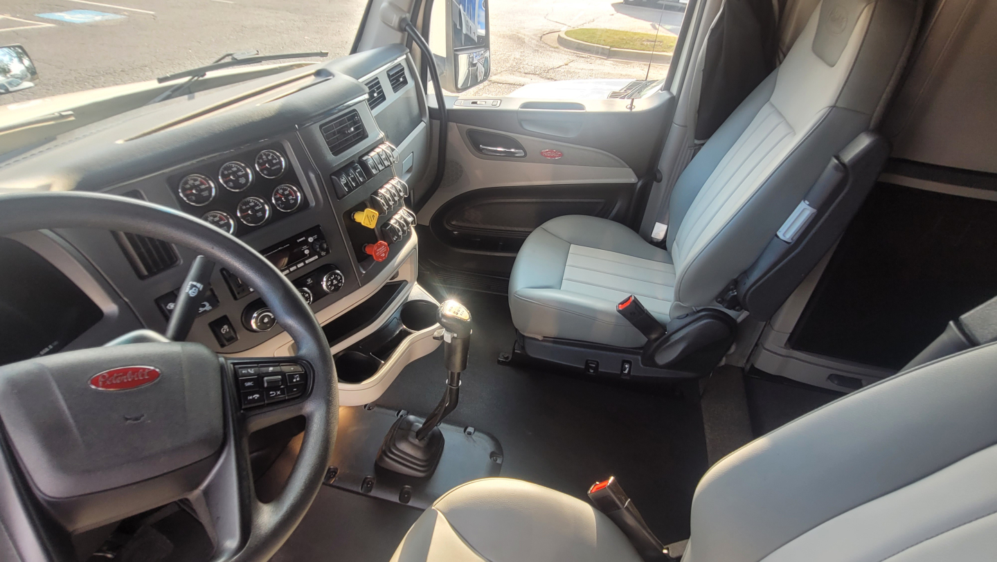 Truck cab interior of a 2025 Peterbilt 579 with steering wheel, dashboard gauges, and two light-gray seats seen from the driver side.