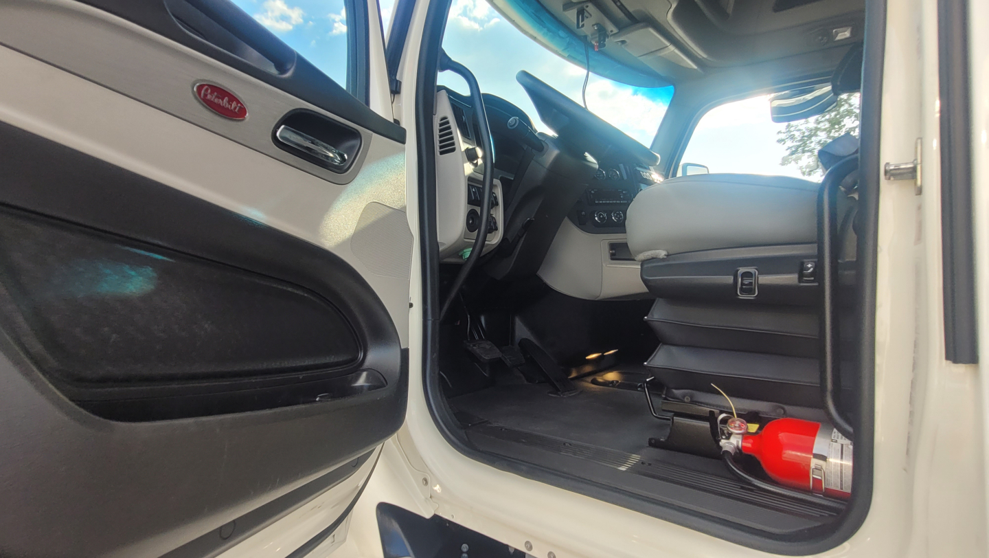 Peterbilt 579 cab interior showing open driver door, steering wheel, dashboard, and a red fire extinguisher on the floor near the seat.