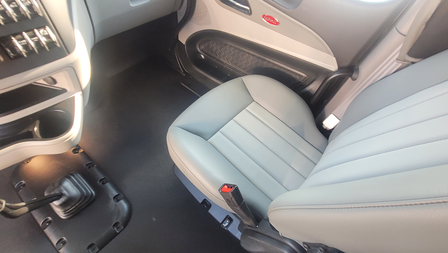 Interior view of a Peterbilt truck cab showing the driver seat and passenger seat with light gray upholstery, floor panels, and part of the dashboard; no specific model text visible.