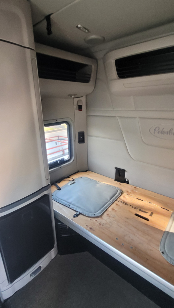 Peterbilt 579 truck cabin interior showing sleeper bunk with wooden bed surface and grey cushions, window hatch and white panel walls.