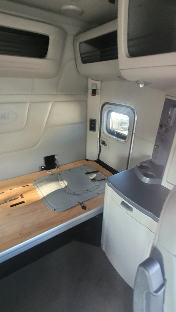 Peterbilt 579 cab interior showing sleeper area with bed platform and window, storage cabinets above, and light gray panels.