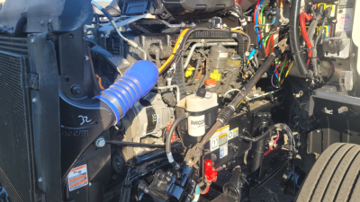 Peterbilt 579 engine bay showing various components, wiring, and fluid reservoirs including a blue silicone hose, power steering fluid bottle, and battery area.
