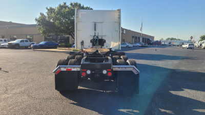 Peterbilt 579 rear view showing fifth wheel, rear tires, and chassis with trailer hookup; no cab in view; parking lot with other vehicles in background.