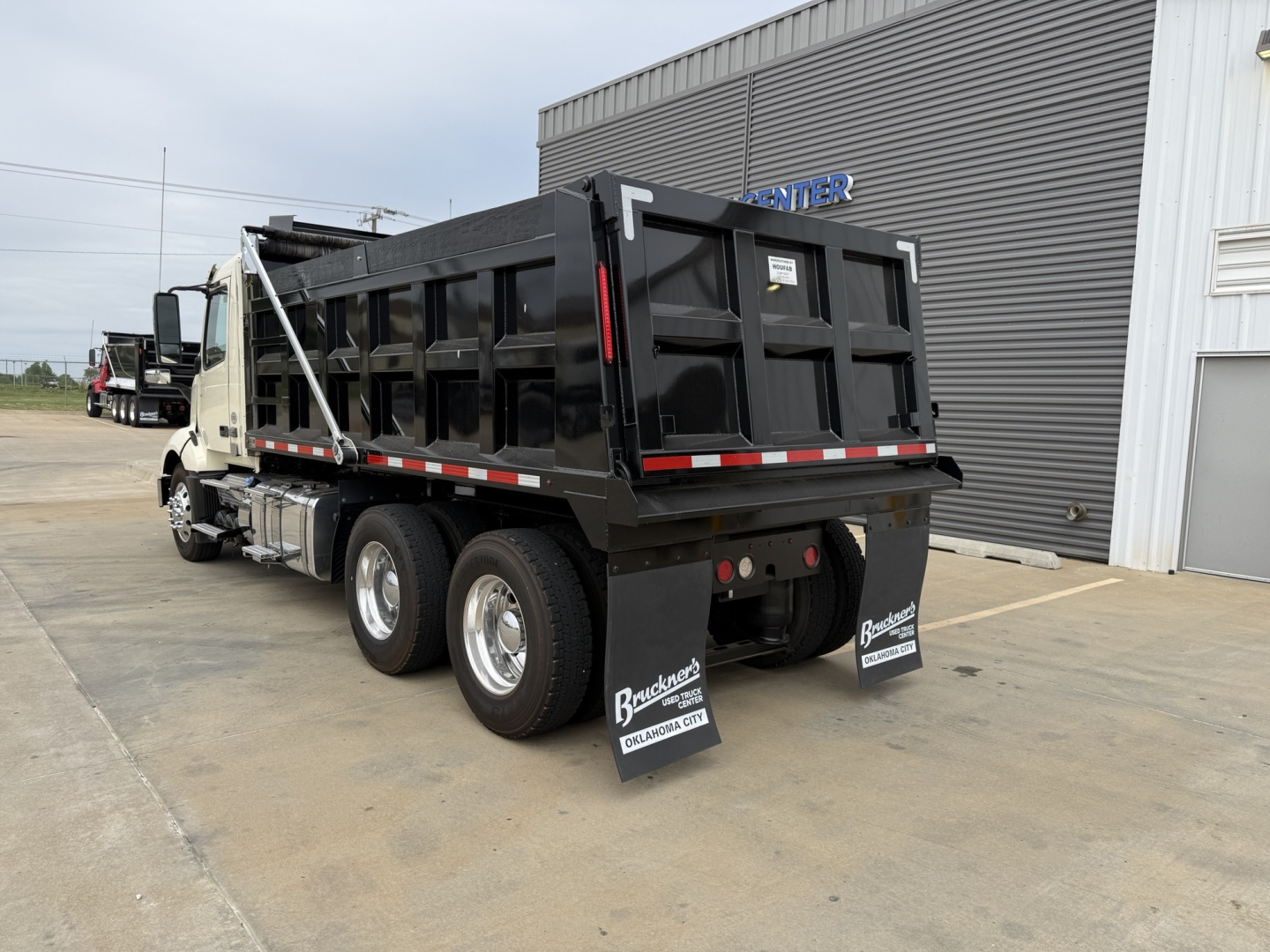 Dump truck rear and side view showing cargo bed with black sides, rear wheels, mud flaps with branding, and a Volvo VNL300 cabin in distance. Visible text includes Bracewell Trucking and dealership info on mud flaps.