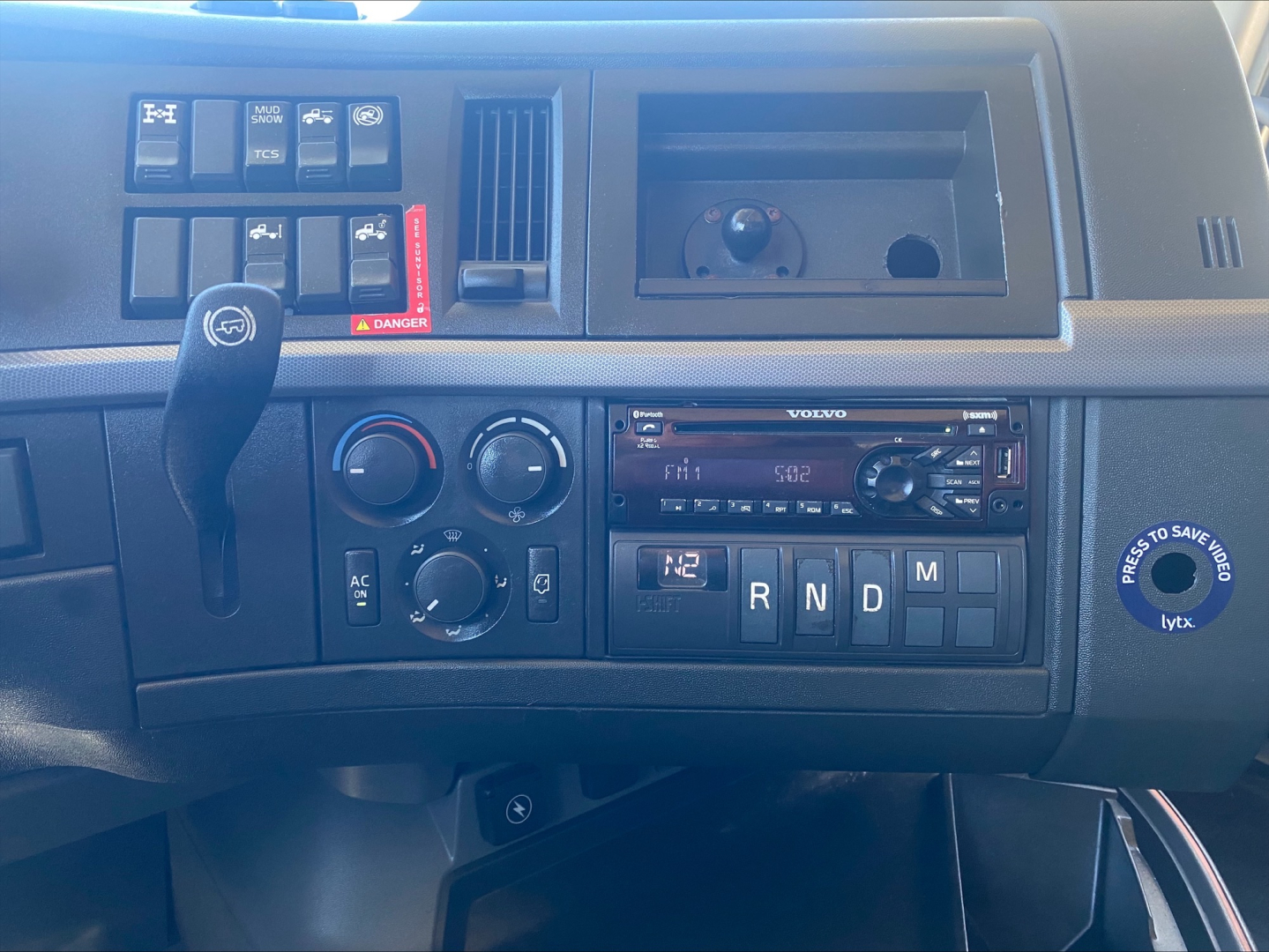 Volvo truck instrument panel showing climate controls, gear selector with R N D M, and radio in-dash, dash storage bin, and various switches.