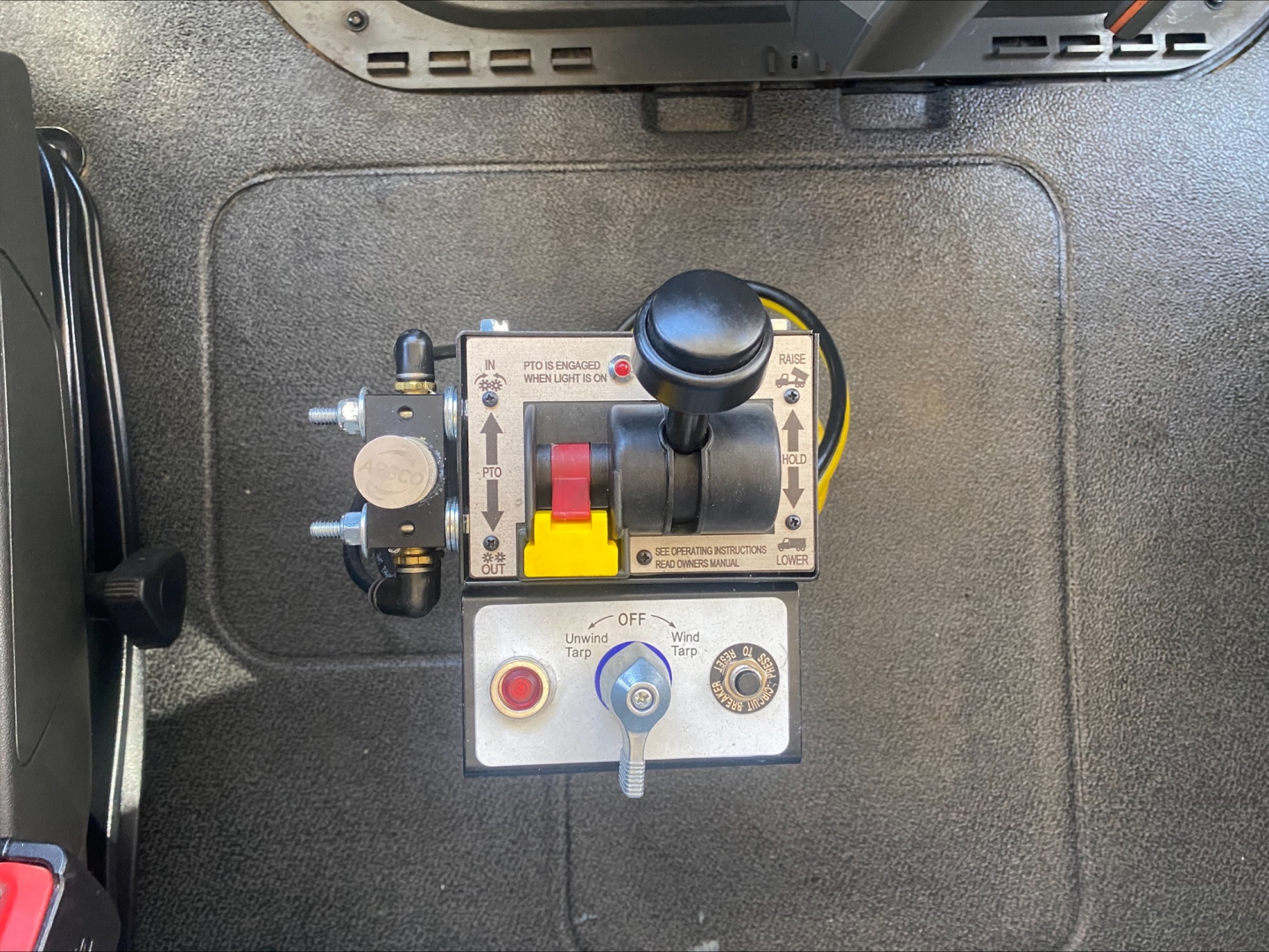 Tractor truck interior showing auxiliary control panel with levers and gauges; circular black knob, red/yellow lever, and warning labels; area appears to be a storage or access panel in a Volvo VNL300 dump truck.