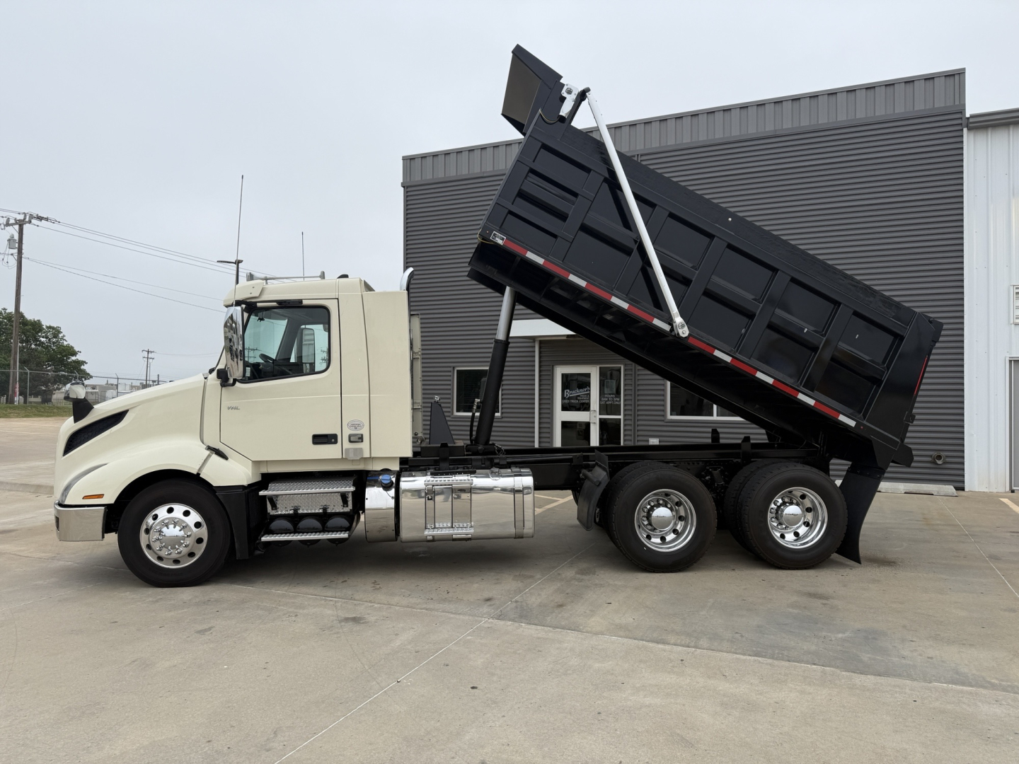 Volvo VNL300 dump truck with bed raised showing side profile and cab. Text on truck includes no visible labels; overall image depicts a beige cab with chrome fuel tanks and a black dump body in raised position.