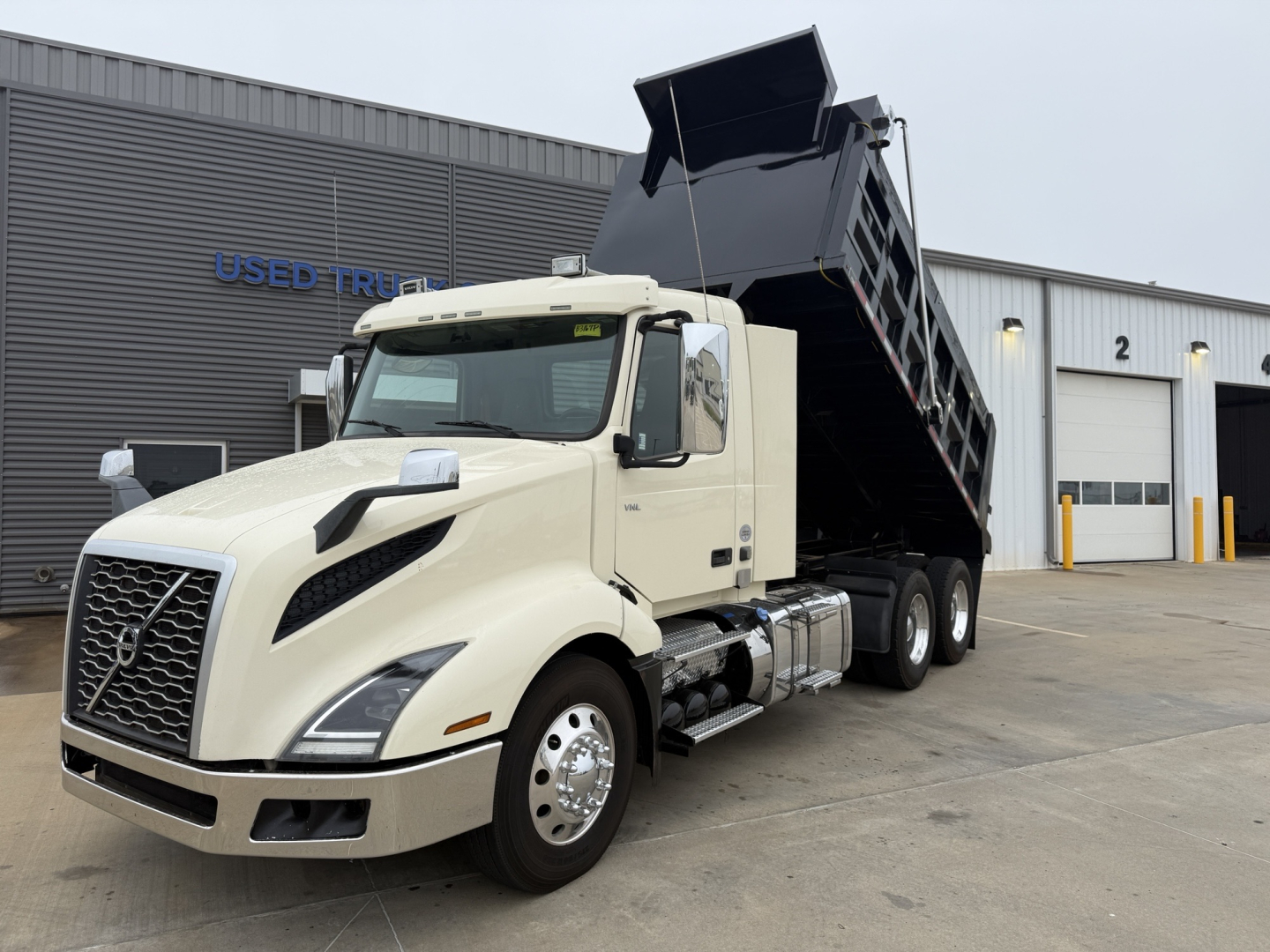Truck in front of a depot with its dump bed raised, 2021 Volvo VNL300 dump truck in cream color, VIN 4V4NC9EG6MN269611 visible on listing text