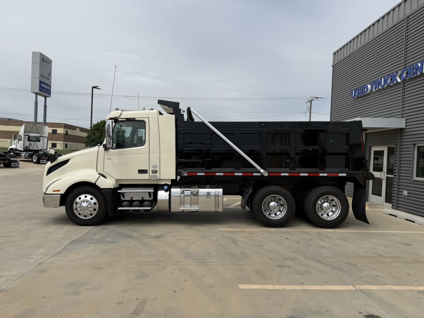 Dump truck side view showing Volvo VNL300 with black dump bed and chrome fuel tanks. Front cab is white, stock number B3167P visible near dealership building.
