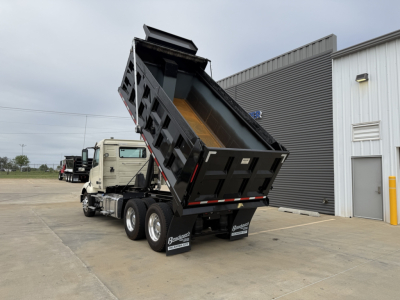 Dump truck with rear dump bed raised, Volvo VNL300 chassis visible, black dump bed, dual rear axles, lot with a building in background.