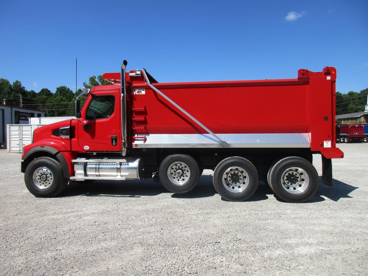 Western Star 49X dump truck side view showing red dump body, chrome fuel tanks, and triple rear axles in a gravel lot.