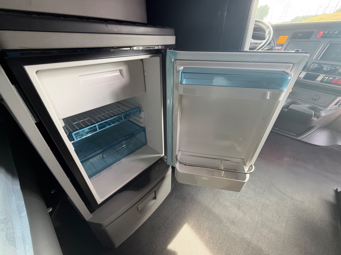 Kenworth T680 cabin interior showcasing open under-dash refrigerator with small freezer and door shelves, blue tray, and cockpit visible.