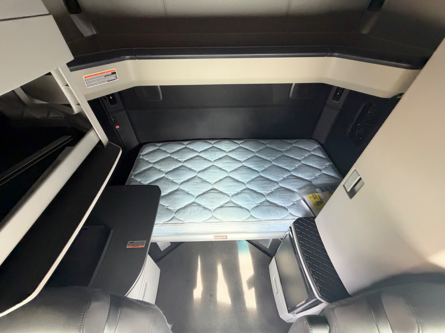 Kenworth T680 sleeper cab interior showing diamond quilted sleeper mattress and driver-side steps; storage compartments and dash area partially visible.