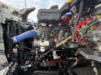 Engine bay of a Kenworth T680 with exposed components, hoses, and wiring; visible are a Packair or PACCAR engine cover, coolant reservoir, various fluid lines, and electrical harnesses.