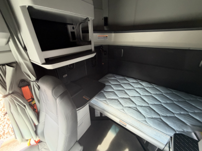 Kenworth T680 sleeper cab interior showing bunk bed with light blue quilted mattress, storage compartments, and driver seat in view.