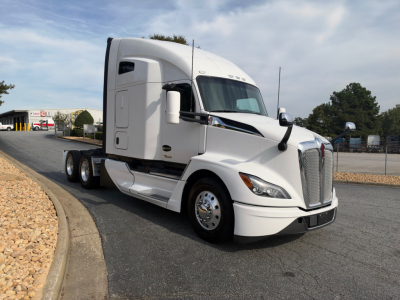 2023 Kenworth T680 tractor in a parking lot, front three-quarter view, white paint, sleeper cab, dual rear axles, chrome wheels.