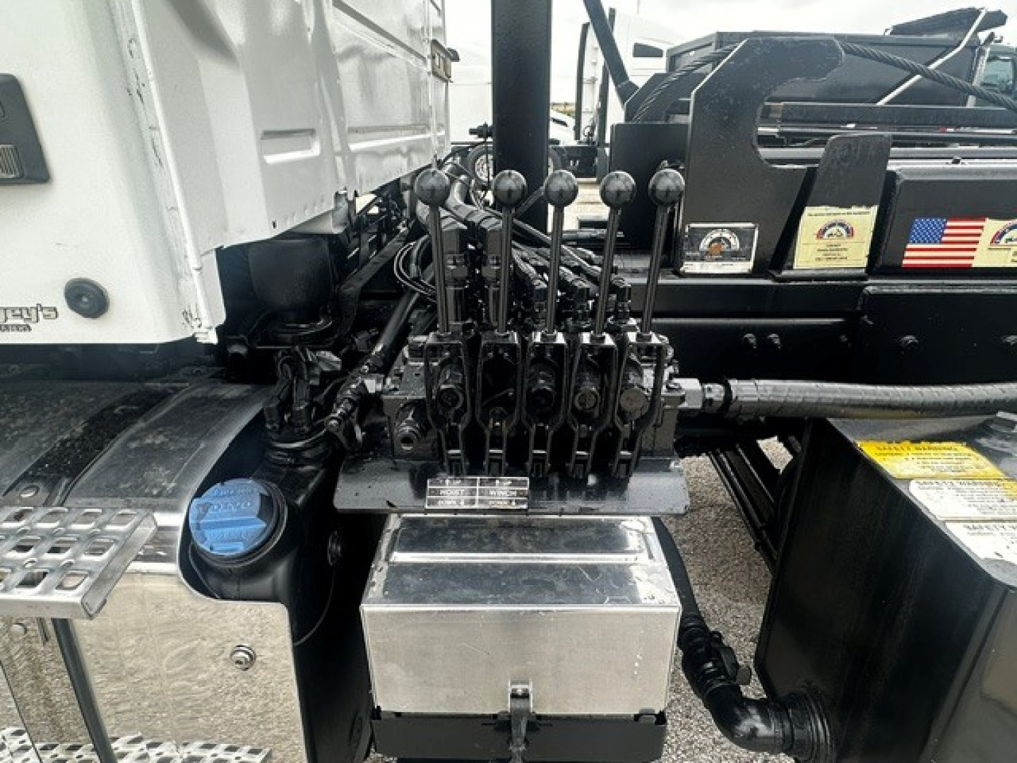 Volvo VHD cab area showing multiple hydraulic levers and chrome toolbox with blue cap, surrounding hoses and metal steps.
