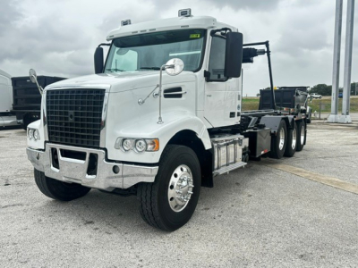 White Volvo VHD truck in a yard showing front grille, cab, and a roll-off flatbed chassis with dual rear axles.