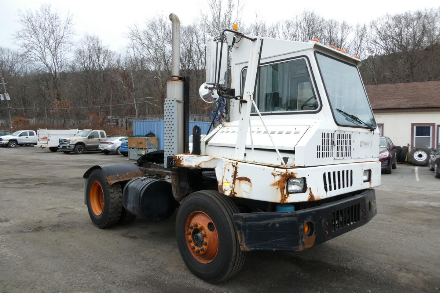 Truck tractor in yard with white cab, upright exhaust stack, and orange wheels; visible rust on frame and metal components.