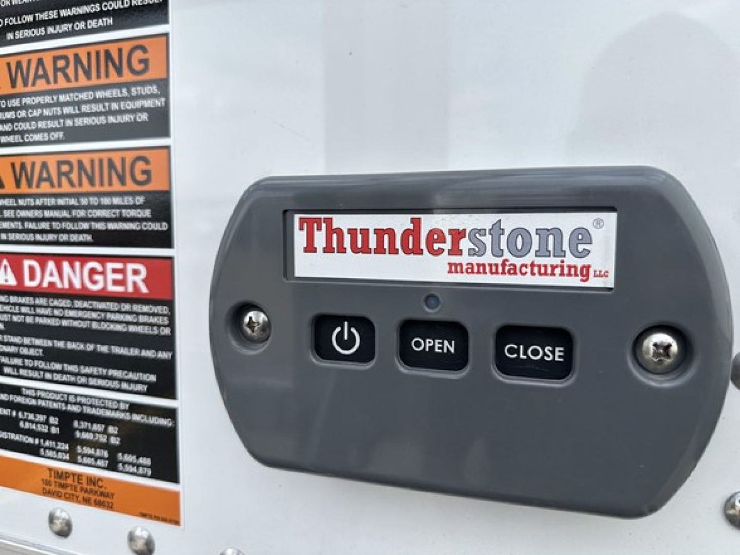 Trailer control panel with Thunderstone manufacturing logo and three buttons labeled OPEN, CLOSE, and power symbol; warning labels on left edge of the frame.