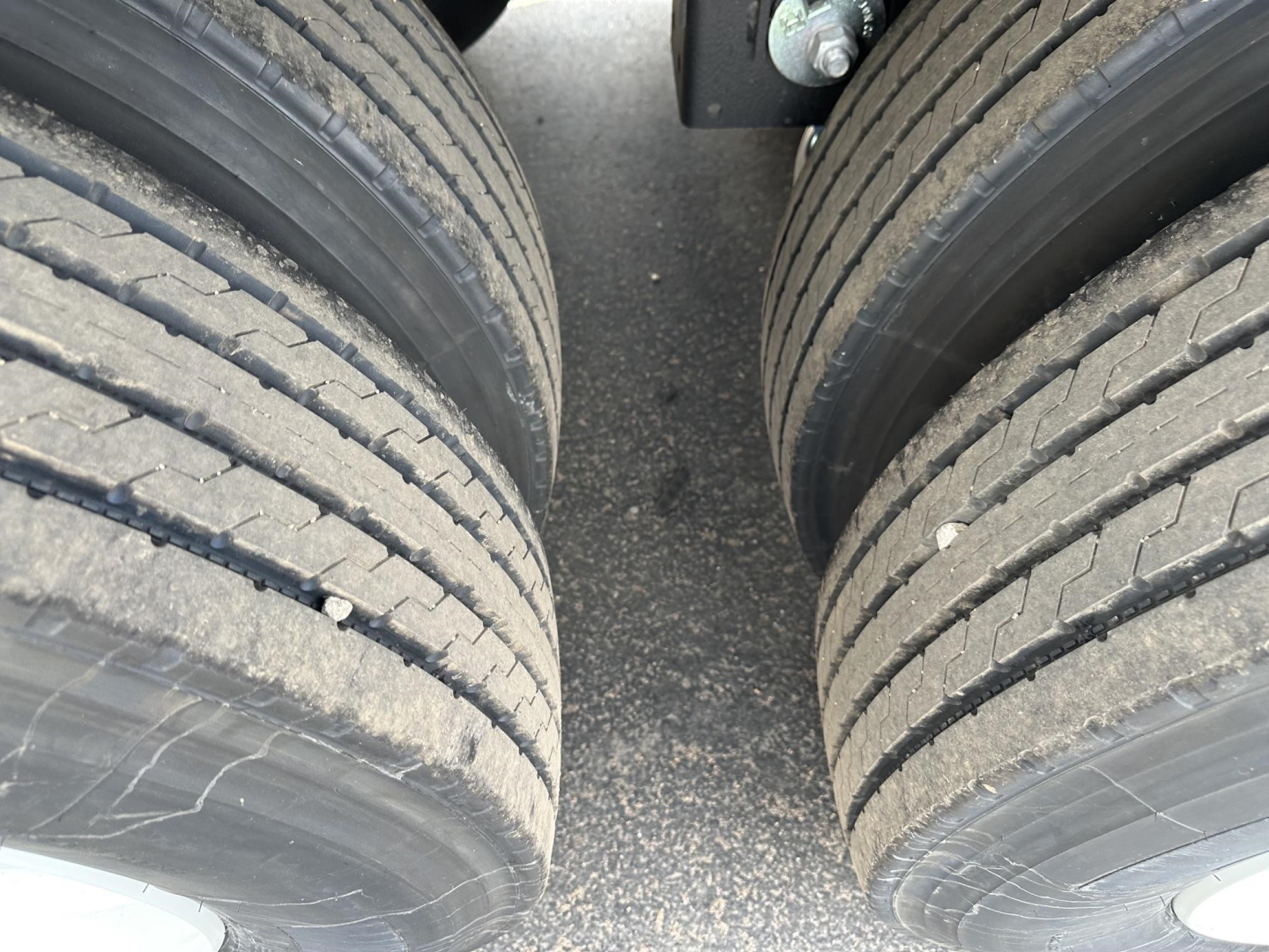 Trailer tires and frame gap between dual axles; close-up of rubber tread on tires and grey asphalt surface between wheels.