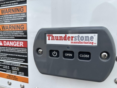 Trailer control panel with Thunderstone manufacturing logo and three buttons labeled OPEN, CLOSE, and power symbol; warning labels on left edge of the frame.