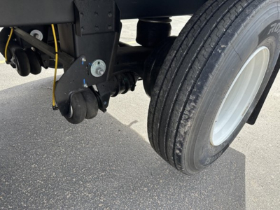 Trailer undercarriage showing suspension components and single wheel with tire, hydraulic hose, and metal brackets.