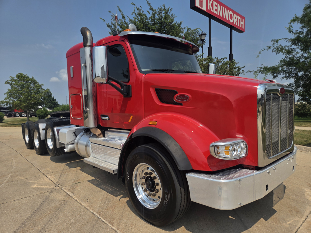 Peterbilt 567 day cab truck in red, front-three-quarter view, chrome grille, dual rear axles, Kenworth dealership sign in background