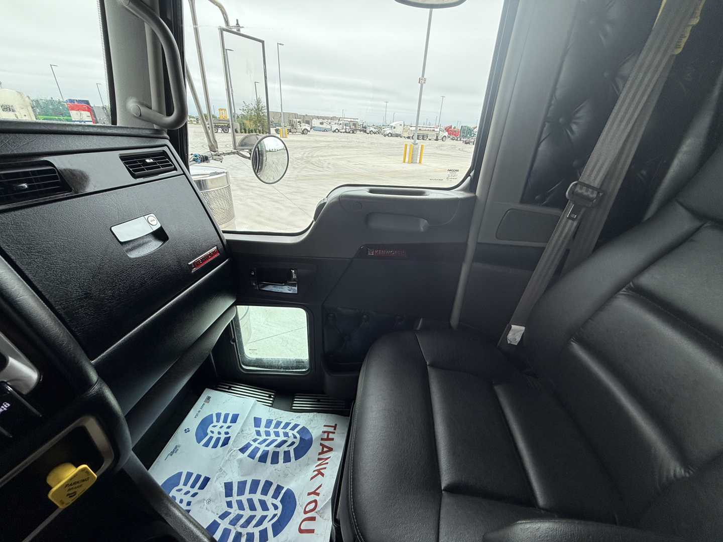 Cab interior of a 2023 Kenworth W900L truck showing front passenger seat, dashboard, door panel, and window with reflection of outside lot.