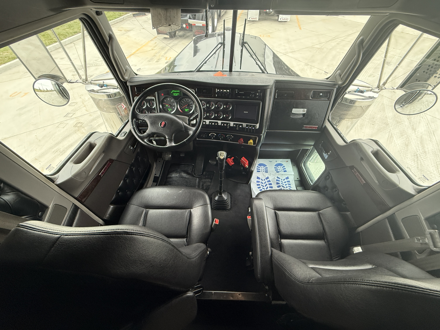 Kenworth W900L cab interior showing steering wheel and dashboard with gauges, gear shifter, and driver/passenger seats.