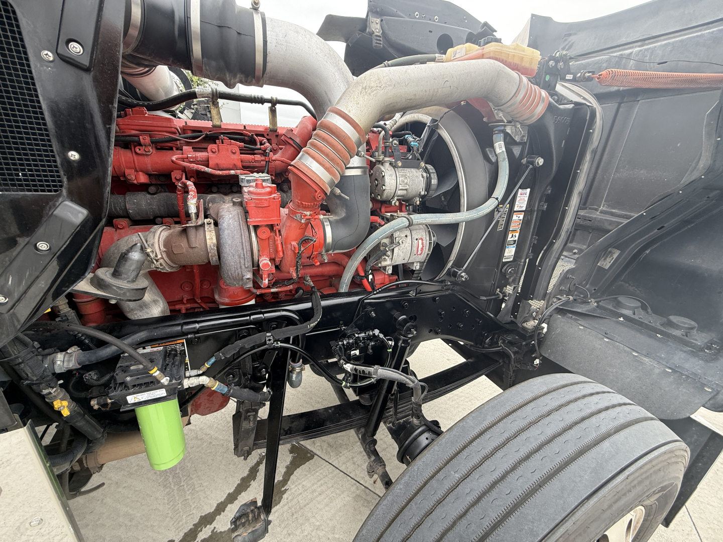 Kenworth W900L engine bay showing red diesel engine with tubing, hoses, and components; green filter canister visible under hood.