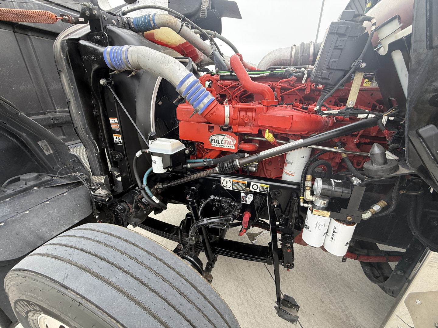 Kenworth W900L engine bay showing red diesel engine and nearby components, hoses, filters, and frame with blue and white hoses visible.
