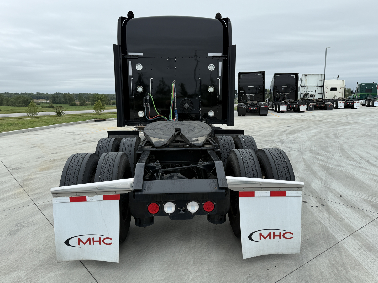 Kenworth W900L rear view showing exhausts, dual rear axles, mud flaps with MHC logos, and wiring harnesses; no visible model text other than the truck components.